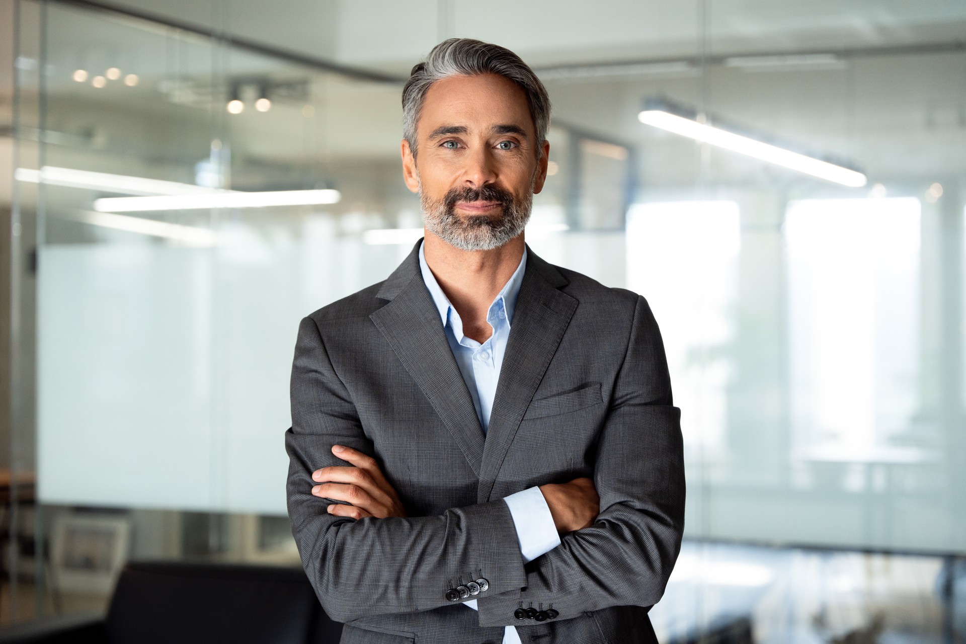 Handsome hispanic senior business man with crossed arms smiling at camera. Indian or latin confident mature good looking middle age leader male businessman on blur office background with copy space.